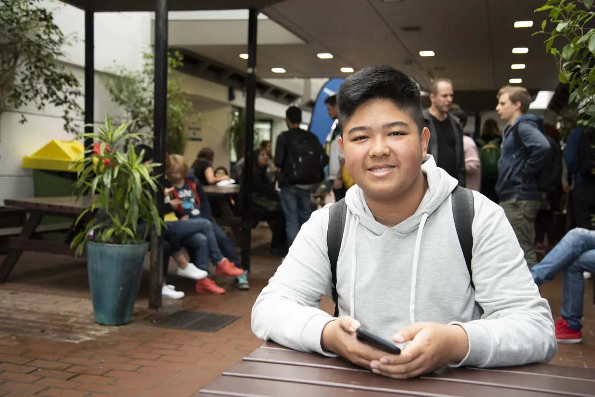A student with a phone at an outdoor table in the busy campus courtyard