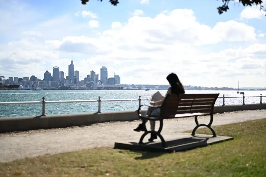 A person reading on a bench at the Devonport waterfront, looking across the harbour to the Auckland skyline