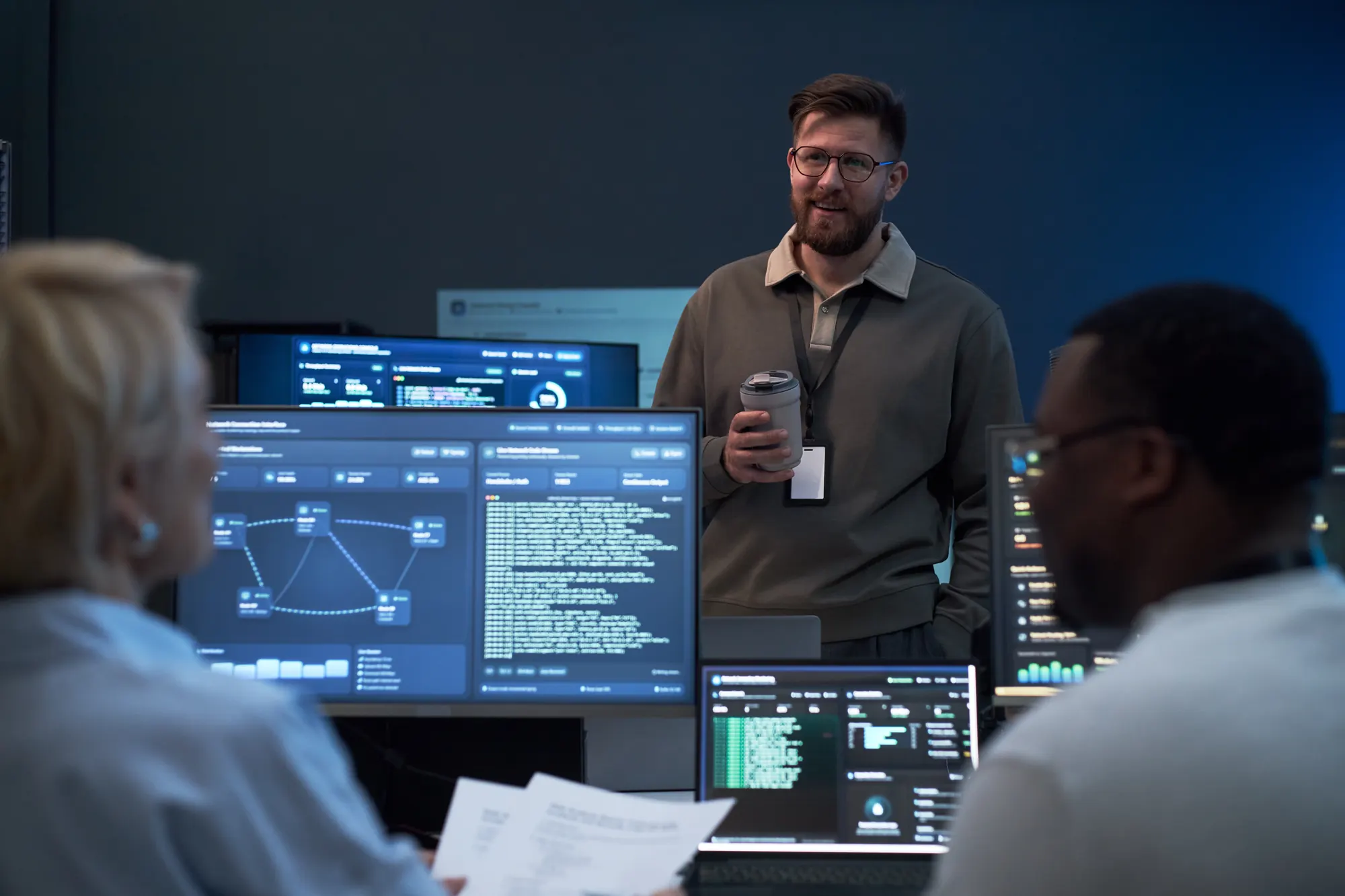 An IT instructor talking with students in a technology lab surrounded by network and code dashboards