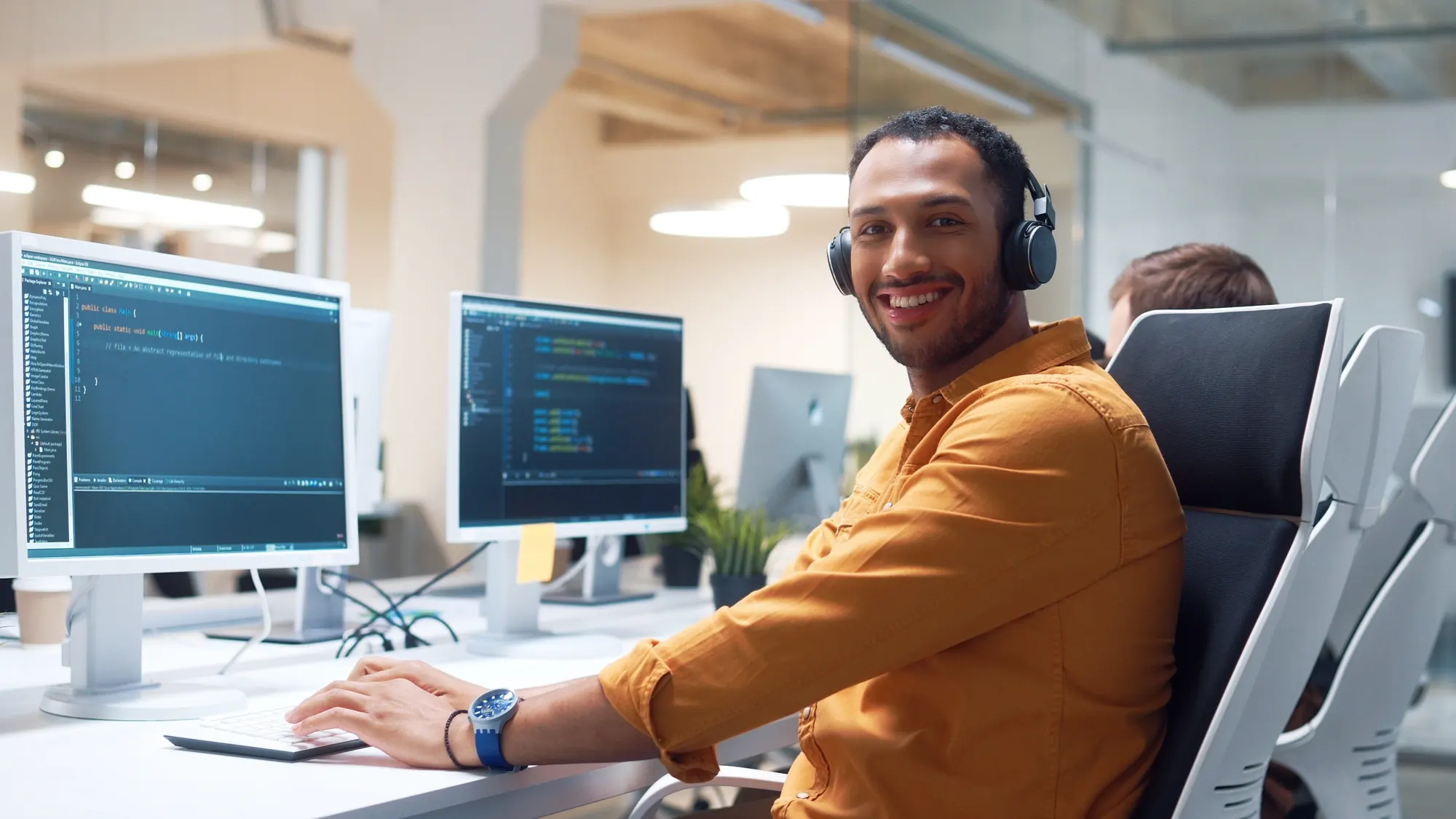 A smiling software development student wearing headphones at a desk with code on two monitors
